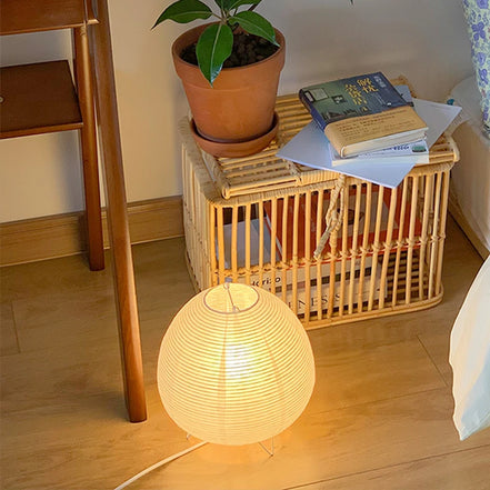 Haru lamp lit on a wooden floor beside a rattan side table and plant